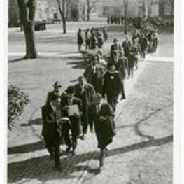 A group of men walk down a brick sidewalk with college buildings in the background.