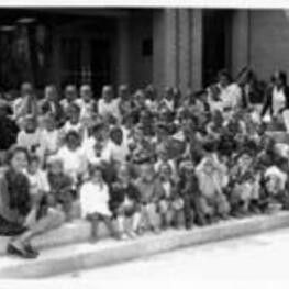 A large group of unidentified children sit on steps outside of a building and smile.