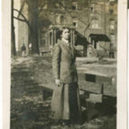 A woman poses next to a bench in front of a large building.