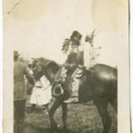 Two young men dressed as Native American caricatures sit on a horse being led by another man.