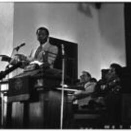 A man addresses people in a church, while Maynard Jackson sits in the background.
