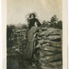 Two women look at each other. One woman sits on a pile of logs while the other stands next to the logpile.