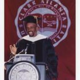 A man, wearing graduation cap and gown, stands at the podium at commencement in front of a banner "Clark Atlanta University."