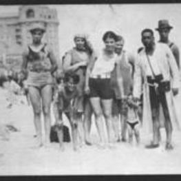 A group of men, women, and children standing on a beach with a large building in the background.