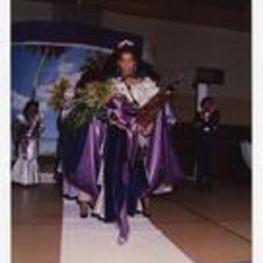 View of woman holding boquet of flowers and award trophy walking on stage. Written on verso: "Miss Morris Brown College 1987-88 Julliette Burgess".