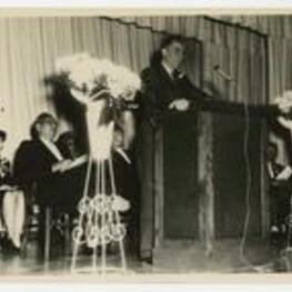 Dr. William Gill speaks at a podium with men and women on stage. Written on verso: Dr. William Gill, Vice President, Mississippi State University, Centennial Convocation Speaker, February 11, 1962, female: Alcon oldest grad.