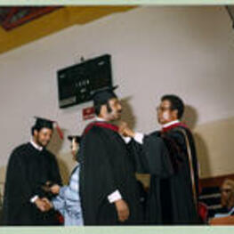 An unidentified graduate is given a red hood by a faculty member.