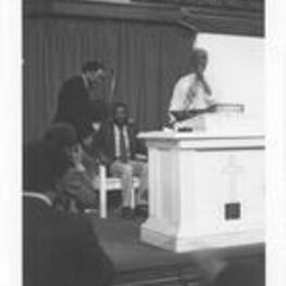An unidentified man speaks at a lectern while Dick Gregory sits.