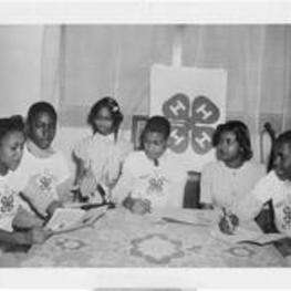 A group of young boys and girls wearing 4-H t-shirts study at a table. Written on verso: 4-H Boys and girls.