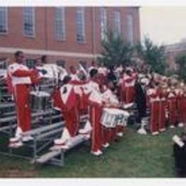 Men and women, wearing red and white marching band uniforms, play musical instruments while standing on bleachers outdoors at convocation.