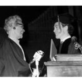 Grace Towns Hamilton stands at a podium shaking hands with a man wearing a cap and gown. Written on verso: John Spencer? A.U. Ch. [illegible] 9. T.H-Oct. 16-1981.