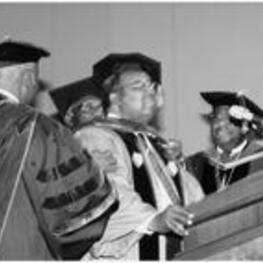 C. Eric Lincoln, wearing regalia, stands at a podium and receives a hood in a ceremony.