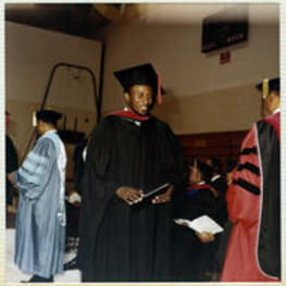 An unidentified man in his cap and gown smiles while holding his diploma.