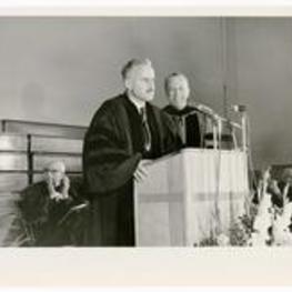 An unidentified man speaks at a podium while Hugh Morris Gloster looks on.