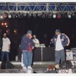 Two men, wearing athletic jackets and baseball caps, hold microphones on stage, a disc jockey and other men stand in the background.