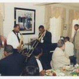 Wynton Marsalis plays his trumpet with a band of four men, playing saxophone, piano, upright bass, and a trombone, in a dining room, while men and women sit at dining tables.