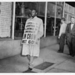A man walks in front of a store wearing a sign that reads, "The presence of segregation is the absence of democracy, Jim Crow must go!".