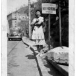 An unidentified woman stands on the curb of a town street. She stands next to a sign reading: "Positively No Parking. 10.00 fine." A car is parallel parked behind her.