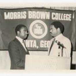 Two men at a podium, flag in background: Morris Brown College. Written on verso: "L to R; Head football coach- Gregory Thompson; Head Basketball Coach- Harold Merrill".