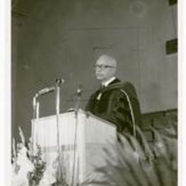 An unidentified man speaks at a podium wearing a commencement robe.