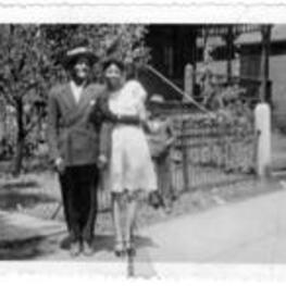An unidentified couple stand on the sidewalk in front of an large house with an iron fence around the front yard. An unidentified boy is standing behind them, wearing a suit and a hat.
