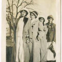 A group of four women stand outside with hats and long coats.