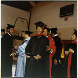 An unidentified man smiles as he holds his diploma. An unidentified faculty member stands behind him.