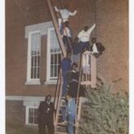 Outdoor group portrait of young men and women on fire-escape ladder. Written on verso: "1986 Brownite Yrbk Staff; From Top to bottom- Phillip Tate, Yvette Paris, Todd Blackburn, Travis Goodson, Shawnee Jackson, Michael Childress, John Jackson, Guinee Cook, Vincent E. Harris, Editor in Chief, Mr. Charles Barker, Consultant".
