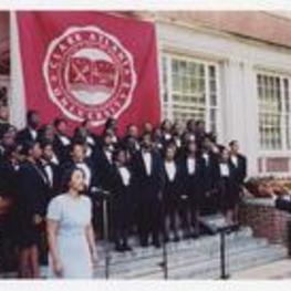 A group of men and women, wearing suits with bow ties, sing on steps of a building under a banner "Clark Atlanta University."