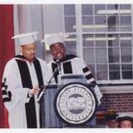 Two men, wearing graduation cap and gowns, stand at the podium at commencement.