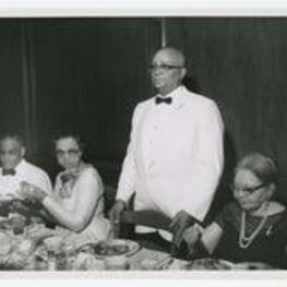 Sidney Williams stands at a banquet table with others seated. Written on verso: Sidney class of 14, Mr. and Mrs. Williams.