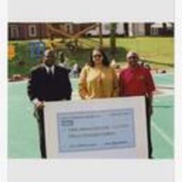 Dr. Walter Boradnax poses with another man and a woman holding a large check "Imiren Pharmaceuticals, Inc., September 2002, Pay to Clark Atlanta University $15,000," on football field.