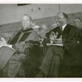 Hugh Morris Gloster and an unidentified man sit on stage and clap.