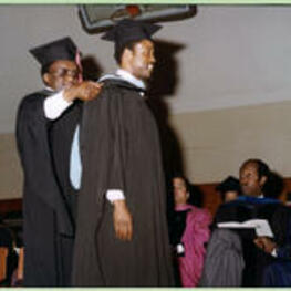 An unidentified faculty member places a blue and black hood on an unidentified graduate.