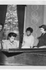 A woman sits at the piano and group of women stand around the piano. Written on verso: Dormitory life- November 1952. Students relaxing in Bumstead Hall
