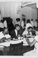 An unidentified group eats at the table while others stand in line. Written on verso: Area Pastor's School at Gammon.