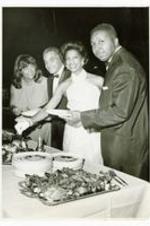View of two men and two women at buffet table with platters of food and plates. Written on verso: l to r, Mrs. Richard Handy, Myron Johnson, Mrs. Myron Johnson, Dr. Richard Handy