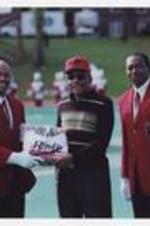 Dr. Walter Boradnax poses with two other men, wearing matching red jackets and white gloves, on football field.