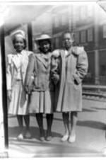 Three unidentified young women stand together and hold hands, possibly at a train station platform.