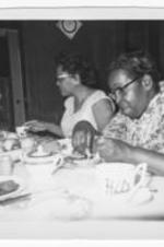 Two unidentified women sit at a dining table together, eating and drinking.