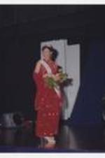 A women, wearing a floor-length red dress with tiara and sash, stands on stage holding a bouquet of flowers.