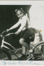A girl rides on a bicycle in front of a house. Written on recto: Jose Anita.