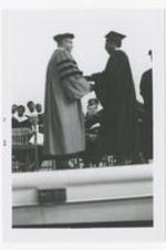 Two men, wearing graduation caps and gowns, shake hands on stage at commencement.