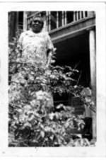 An unidentified woman stands in front of a two-story house house. A second unidentified woman stands on the porch of the first floor.