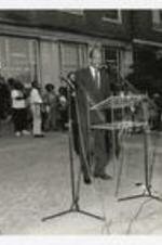 A man stands at the podium in front of a building at a homecoming activity.