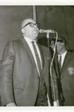 An unidentified man speaks at a microphone with young men wearing 'Morehouse' blazers in background.