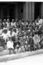 A large group of unidentified children sit on steps outside of a building and smile.