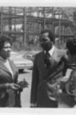 Two unidentified women speak with a man in a parking lot with a building under construction in the background.