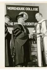 Hugh Morris Gloster shakes hands with a man wearing regalia. On sign in background-Morehouse College, The Second Century.