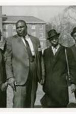 Group portrait of four men wearing suits, with a building in background